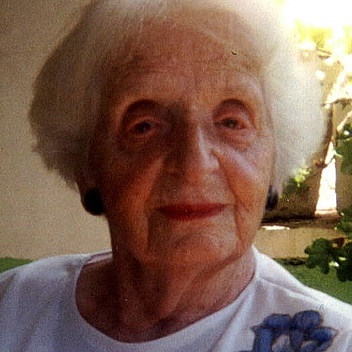 Gerda Wolfsohn-Zondek in einem Zimmer am Fenster sitzend. Sie schaut in die Kamera und trägt einen weißen Pullover mit blauer Blume. Ihr Haar ist kurz und weiß.