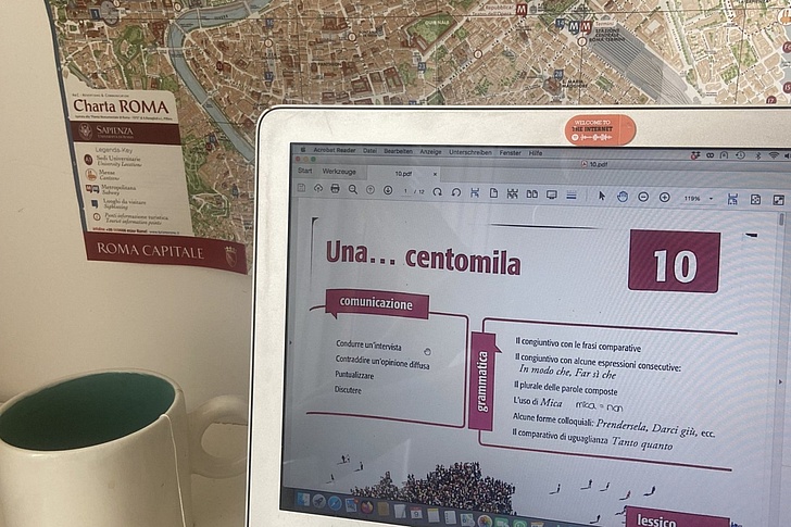 MacBook-Bildschirm zeigt Italienisch-Lektion „Una centomila“ (Kapitel 10) mit Karte von Rom im Hintergrund, daneben weiße Tasse und Teesäckchen auf hellem Tisch.