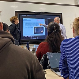 Mehrere Personen sitzen in einem Klassenraum, zwei Personen stehen vor einem großen Bildschirm mit einer Präsentation.