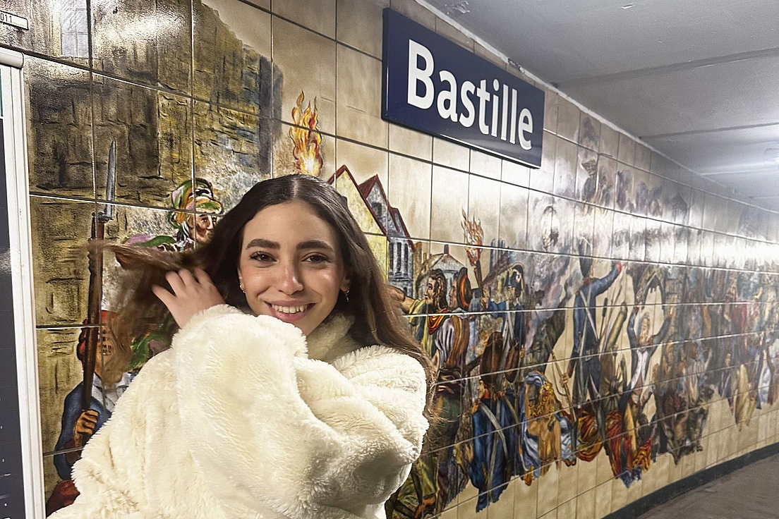 Frau mit langem Haar und weißem Fellmantel vor einer Wand mit einem Wandbild, das die Erstürmung der Bastille zeigt, und einem Schild mit der Aufschrift 'Bastille'