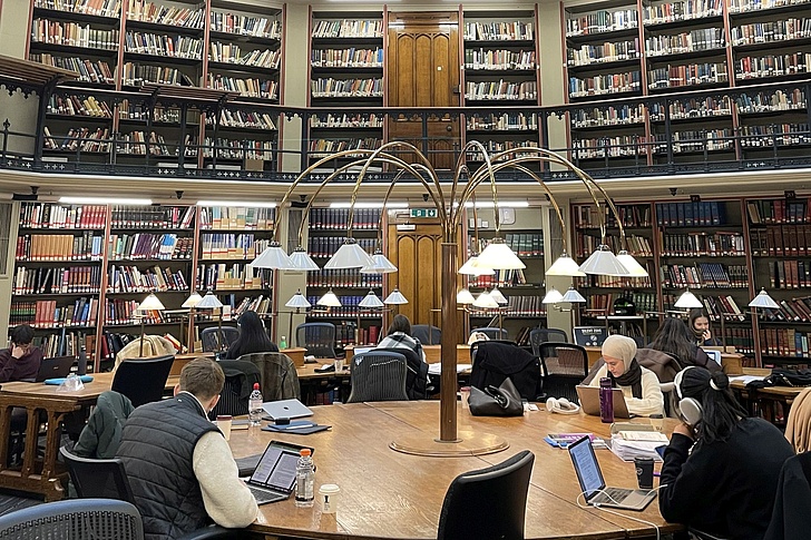 Lesesaal einer Bibliothek mit hohen Bücherregalen, runden Tischen und mehreren Personen, die an Laptops und Büchern arbeiten