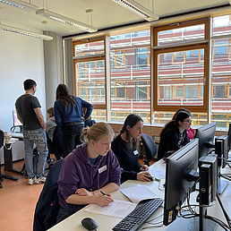 Mehrere Personen sitzen an Computern in einem Klassenzimmer mit großen Fenstern.