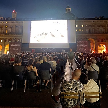 Große Menschenmenge sitzt auf Stühlen vor einer großen Leinwand im Freien bei Nacht in einem historischen Platz mit beleuchteten Arkaden.