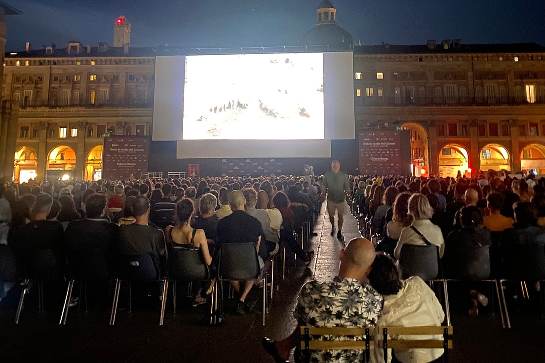 Große Menschenmenge sitzt auf Stühlen vor einer großen Leinwand im Freien bei Nacht in einem historischen Platz mit beleuchteten Arkaden.