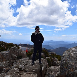 Person steht auf felsigem Gipfel mit Bergpanorama und Wolkenhimmel im Hintergrund
