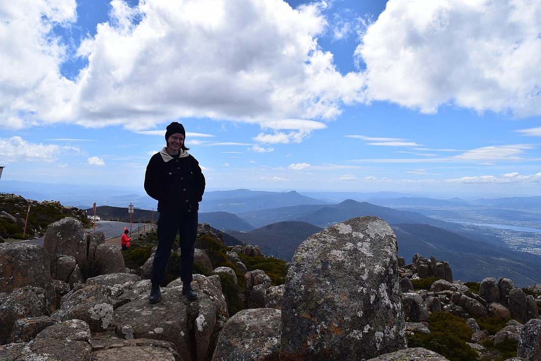 Person steht auf felsigem Gipfel mit Bergpanorama und Wolkenhimmel im Hintergrund