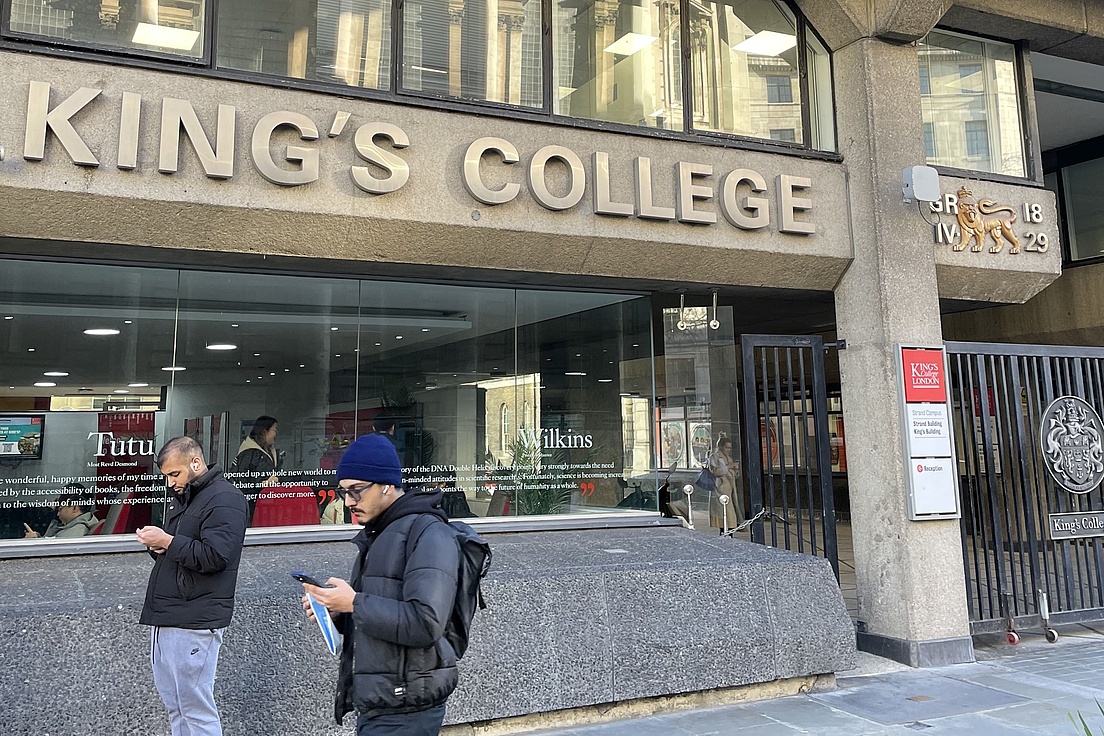 Fassade von King's College mit großen Fenstern und zwei Personen davor, eine davon mit Kapuze und Rucksack, die auf ein Smartphone schaut.