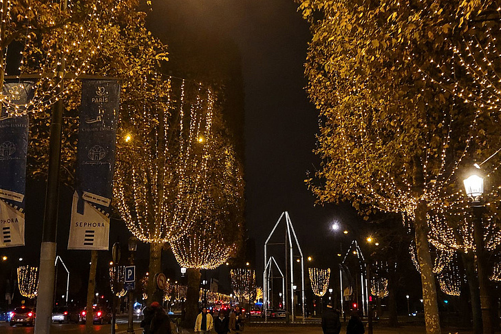 Festlich beleuchteter Stadtplatz nachts mit goldgelben Lichterketten an Laubbäumen, blauen Bannern, weißem Bogen-Tor-Skulptur, Laternen und Passanten bei Autolichtern.