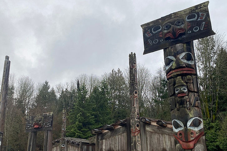 Gruppe traditioneller Totempfähle der First Nations mit geschnitzten Tier- und Ahnenfiguren in Rot, Schwarz und Weiß vor bewaldetem Hügel bei bewölktem Himmel.