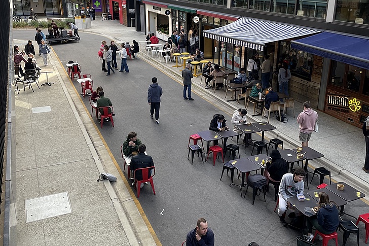 Straßencafé mit mehreren Tischen und Stühlen, an denen Menschen sitzen und stehen, umgeben von Gebäuden mit Geschäften und Markisen.