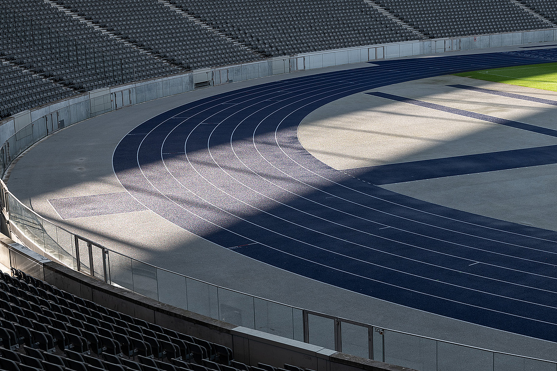 Das Bild zeigt eine leere Laufbahn in einem Stadion. Die Bahn ist blau mit weißen Linien, die die einzelnen Läufe markieren. Im Hintergrund sind Reihen von Sitzplätzen zu sehen, die ebenfalls leer sind.
