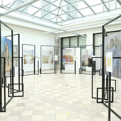 Das Bild zeigt eine Ausstellungshalle mit einem gläsernen Dach, das viel Tageslicht hereinlässt. Der Boden ist mit quadratischen Fliesen in einem Muster aus hellen und dunkleren Farben ausgelegt. In der Halle sind mehrere freistehende, schwarze Metallrahmen aufgestellt, die als Informationspods dienen. An diesen Rahmen sind Poster und Fotos befestigt, die verschiedene Personen und Szenen darstellen.