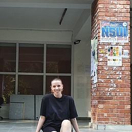 Junge Frau mit Pferdeschwanz in schwarzem T-Shirt sitzt grinsend auf dem Boden vor NSUI-Postern an roten Ziegelsäulen und offenen Fenstern eines Campusgebäudes.