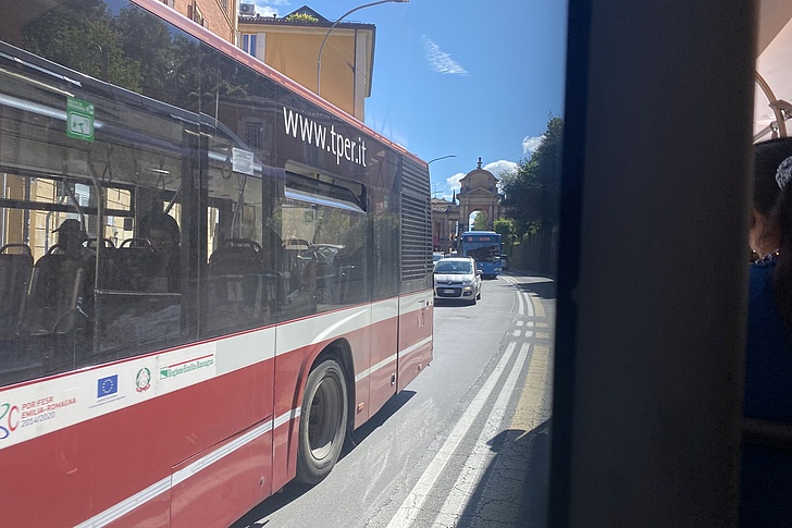 Roter Stadtbus mit der Webadresse 'www.tper.it' fährt auf einer sonnigen Straße mit Gebäuden und parkenden Autos im Hintergrund