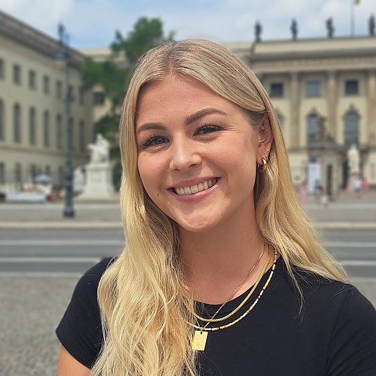 Blonde Frau mit langen Haaren, schwarzem T-Shirt und goldenen Ketten vor einem historischen Gebäude mit Säulen und mehreren Fenstern.
