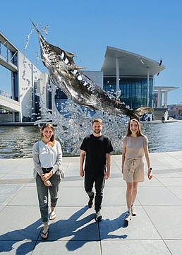 Alicia Burns, Palina Bartashevich und David Mezey stehen vor dem Kampagnenmotiv, auf dem ein gestreifter Marlin abgebildet ist, der aus der Spree springt.