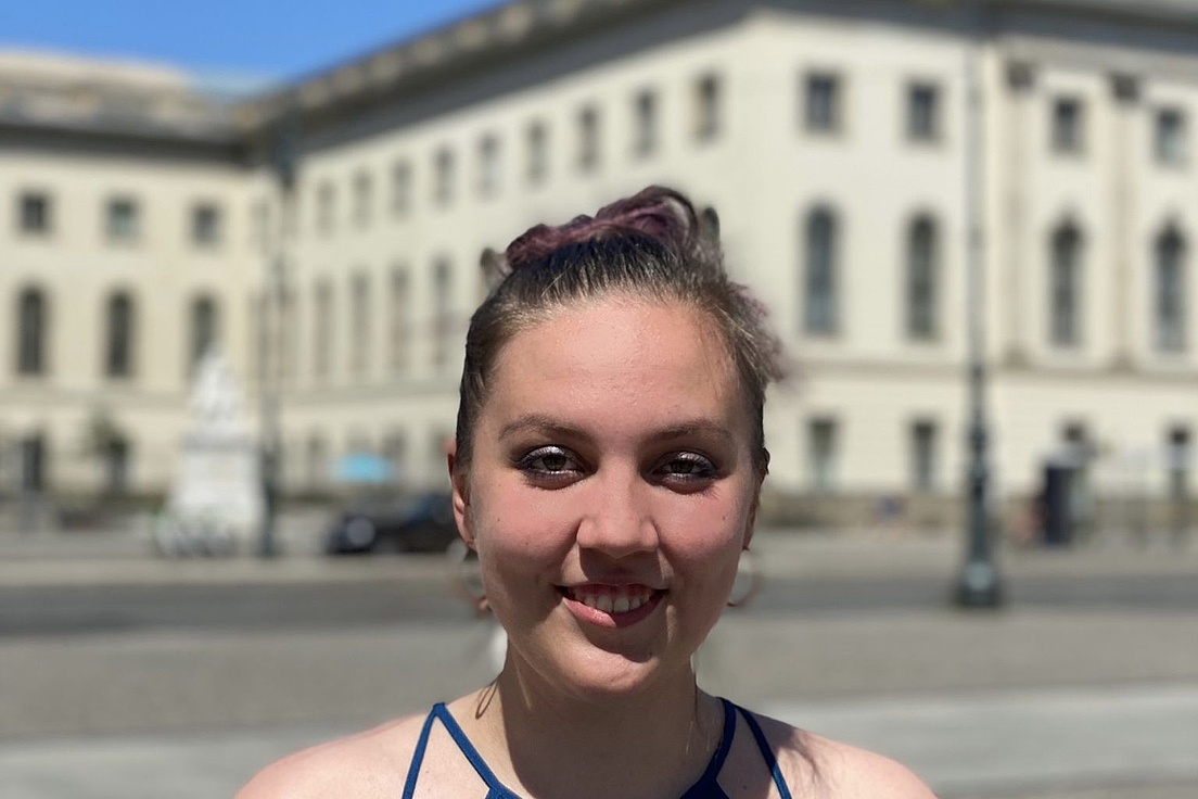 Lächelnde junge Frau mit lila gefärbtem Pferdeschwanz in blauem Top steht vor einem weißen klassischen Gebäude mit Laterne und Skulptur bei blauem Himmel