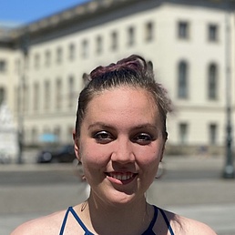Smiling young woman with purple-dyed ponytail in blue top stands in front of a white classical building with lamppost and statue under blue sky.