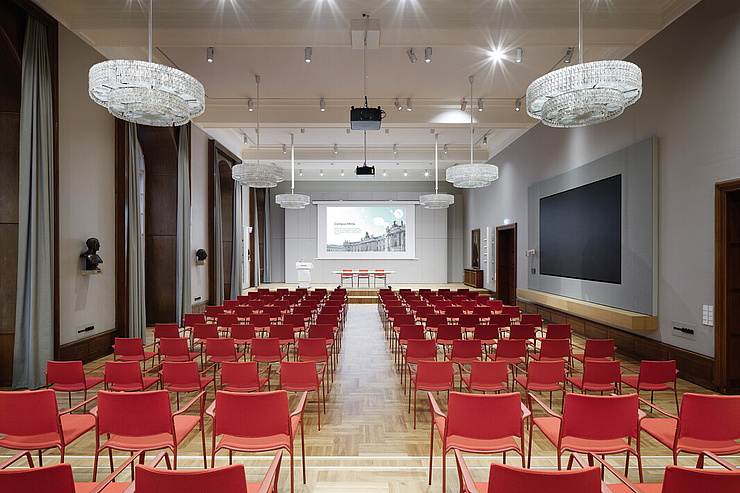 Das Bild zeigt einen großen, eleganten Konferenzraum oder Vortragssaal. Der Raum ist mit zahlreichen roten Stühlen ausgestattet, die in Reihen angeordnet sind und auf eine Bühne mit einem großen Bildschirm zugeordnet sind. An der Decke hängen mehrere große, glitzernde Kronleuchter, die den Raum hell und festlich beleuchten. An der rechten Wand befindet sich ein weiterer großer Bildschirm. Die Wände sind in einem neutralen Grau gehalten und mit Holzvertäfelungen verziert.