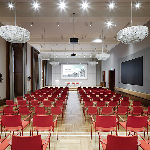 Das Bild zeigt einen großen, eleganten Konferenzraum oder Vortragssaal. Der Raum ist mit zahlreichen roten Stühlen ausgestattet, die in Reihen angeordnet sind und auf eine Bühne mit einem großen Bildschirm zugeordnet sind. An der Decke hängen mehrere große, glitzernde Kronleuchter, die den Raum hell und festlich beleuchten. An der rechten Wand befindet sich ein weiterer großer Bildschirm. Die Wände sind in einem neutralen Grau gehalten und mit Holzvertäfelungen verziert.