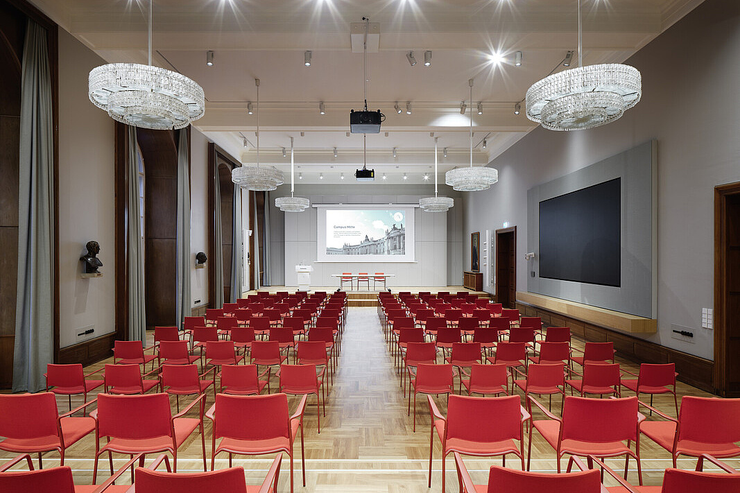 Das Bild zeigt einen großen, eleganten Konferenzraum oder Vortragssaal. Der Raum ist mit zahlreichen roten Stühlen ausgestattet, die in Reihen angeordnet sind und auf eine Bühne mit einem großen Bildschirm zugeordnet sind. An der Decke hängen mehrere große, glitzernde Kronleuchter, die den Raum hell und festlich beleuchten. An der rechten Wand befindet sich ein weiterer großer Bildschirm. Die Wände sind in einem neutralen Grau gehalten und mit Holzvertäfelungen verziert.