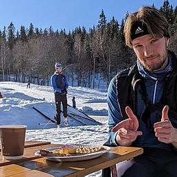 Person in Sportkleidung sitzt an einem Holztisch mit Kaffeebecher und Teller mit Waffel und Kuchenstück im Schnee vor Waldkulisse.