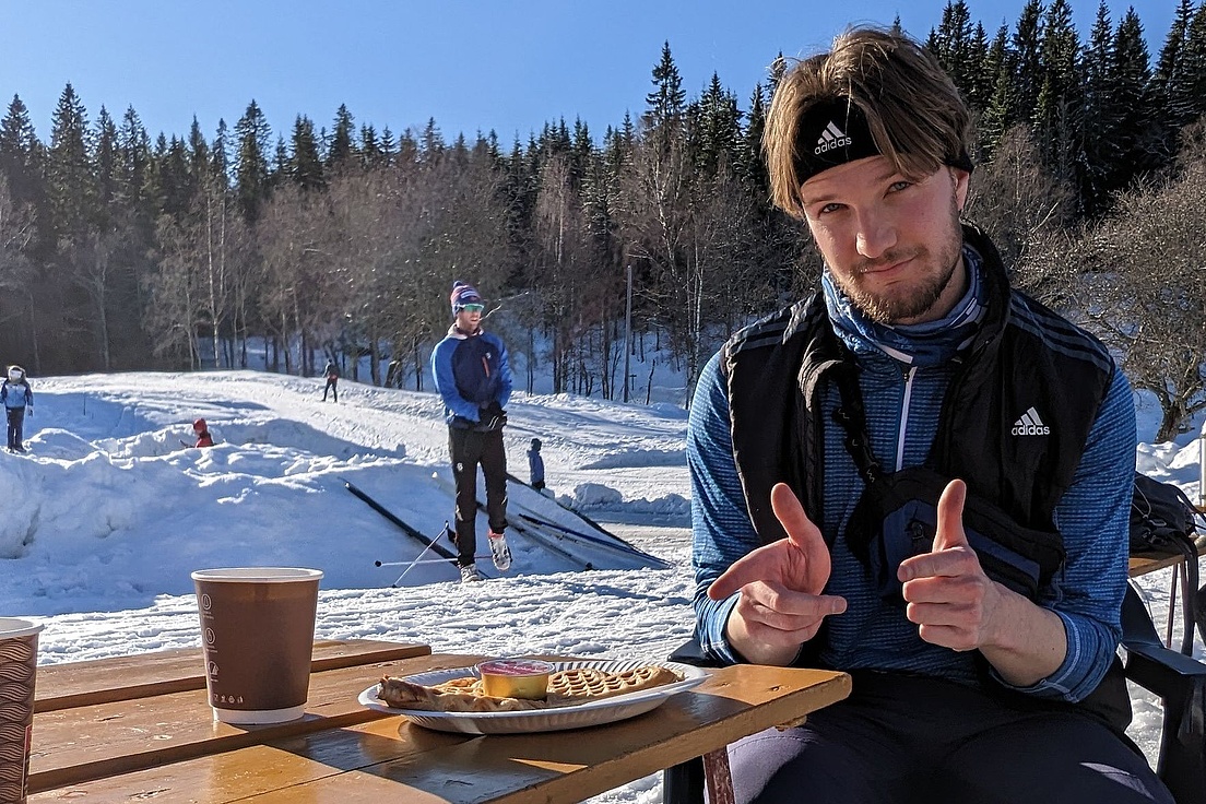 Person in Sportkleidung sitzt an einem Holztisch mit Kaffeebecher und Teller mit Waffel und Kuchenstück im Schnee vor Waldkulisse.
