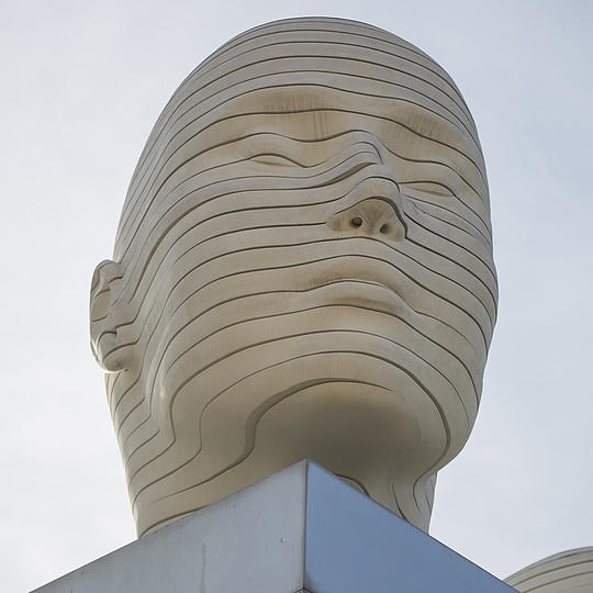 Die Skulptur zeigt einen großen, stilisierten menschlichen Kopf, der auf einem modernen, geometrischen Sockel steht. Der Kopf ist aus einem hellen Material gefertigt, das an Stein oder Beton erinnert, und ist von zahlreichen horizontalen, wellenförmigen Linien durchzogen, die die Gesichtszüge wie Stirn, Augen, Nase und Mund andeuten. Die Linien verlaufen kontinuierlich über die gesamte Oberfläche, wodurch ein fließendes, fast hypnotisches Muster entsteht. Die Augen sind geschlossen.