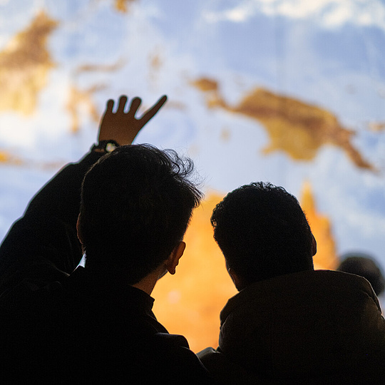 Zwei Menschen erscheinen als Silhouetten vor einer riesigen erleuchteten Erdkugel. Einer von Ihnen zeigt dabei auf die Erde.