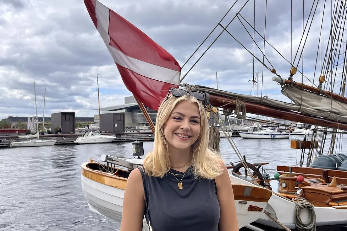 Frau mit blonden Haaren, schwarzem ärmellosem Oberteil und Sonnenbrille auf dem Kopf steht vor einem Segelboot mit einer rot-weißen Flagge im Hafen.