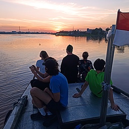 View of a small boat on a lake with young people sitting on it. The sun is setting, the landscape is semi-industrial.