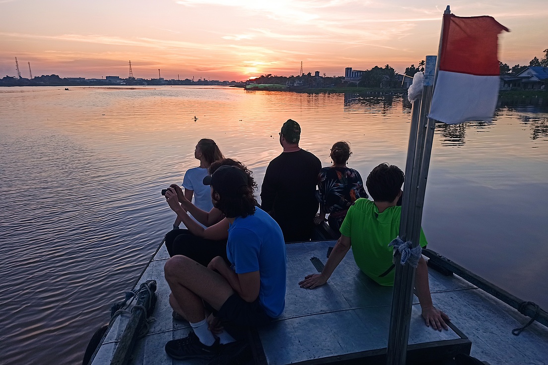  View of a small boat on a lake with young people sitting on it. The sun is setting, the landscape is semi-industrial.