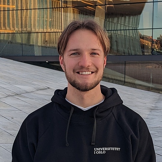 Junger Mann mit schwarzer Kapuzenjacke mit der Aufschrift 'UNIVERSITETET I OSLO' steht vor einem modernen Glasgebäude.