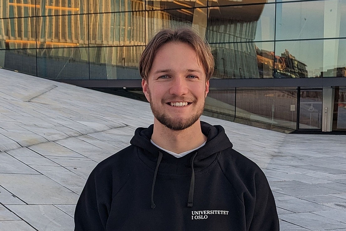 Junger Mann mit schwarzer Kapuzenjacke mit der Aufschrift 'UNIVERSITETET I OSLO' steht vor einem modernen Glasgebäude.
