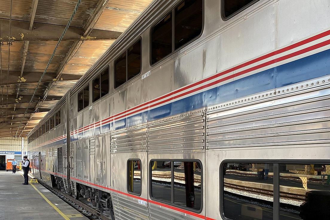 A silver double-decker passenger train is stationed at a covered platform, with a railway staff member in the foreground.