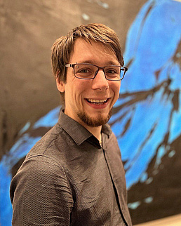 Marco Stucki trägt ein braunes Hemd und lächelt in die Kamera. Er steht vor einem blau-schwarzem Hintergrund