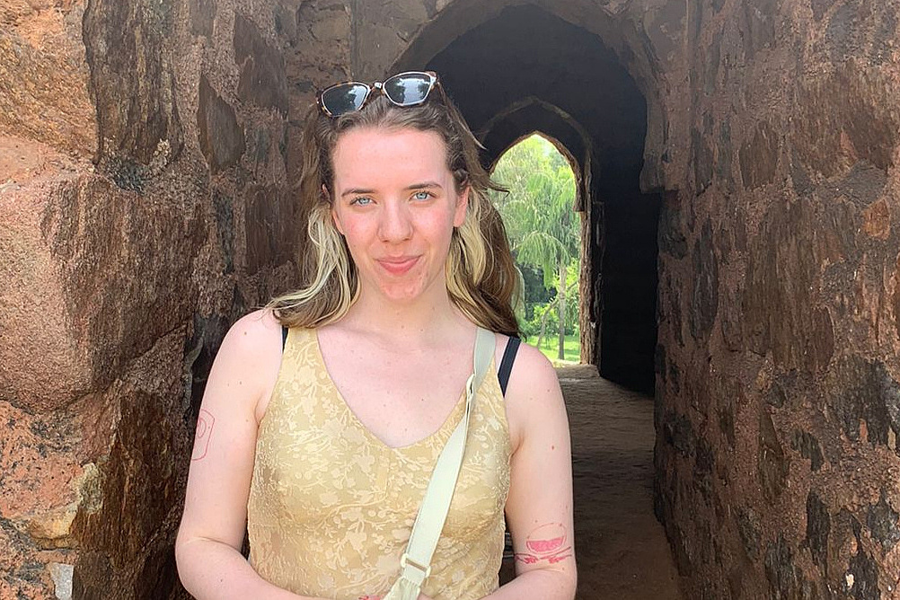 Junge Frau mit hellblondem Haar und Sonnenbrille auf dem Kopf steht lächelnd in einem steinernen Bogen aus rotem Sandstein mit Palmen und Grünanlagen im Hintergrund. Sie trägt ein goldenes Spitzenoberteil und eine beige Umhängetasche.