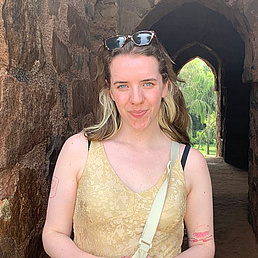 Junge Frau mit hellblondem Haar und Sonnenbrille auf dem Kopf steht lächelnd in einem steinernen Bogen aus rotem Sandstein mit Palmen und Grünanlagen im Hintergrund. Sie trägt ein goldenes Spitzenoberteil und eine beige Umhängetasche.