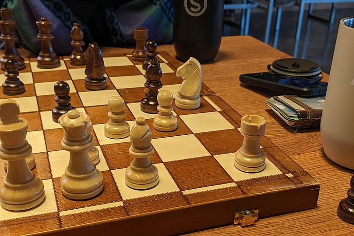 Nahaufnahme eines Schachbretts mit weißen und schwarzen Figuren in einer Spielsituation auf einem Holztisch mit Bechern und anderen Gegenständen im Hintergrund