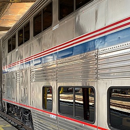 Silberner Doppeldecker-Zug der Amtrak mit roten, weißen und blauen Streifen steht an einem überdachten Bahnsteig, ein Mitarbeiter in Uniform daneben.
