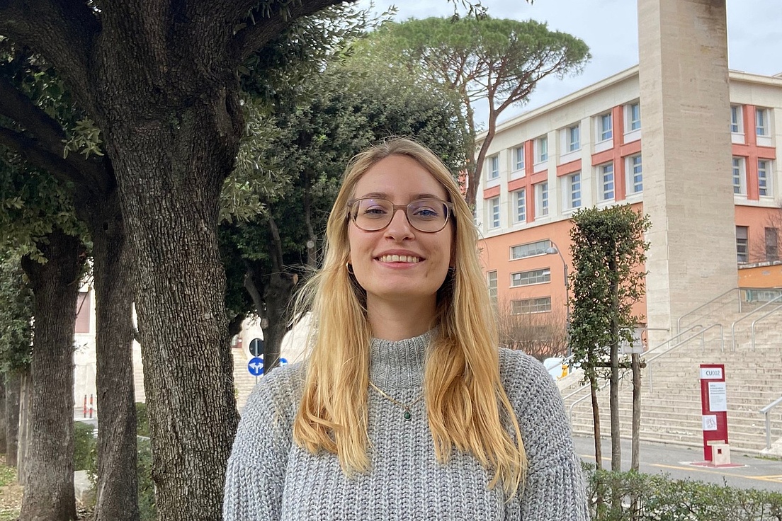 Blonde Frau in grauem Strickpullover steht zwischen schattigen Bäumen vor einem hellen Universitätsgebäude mit breiter Treppe und hohen Säulen.