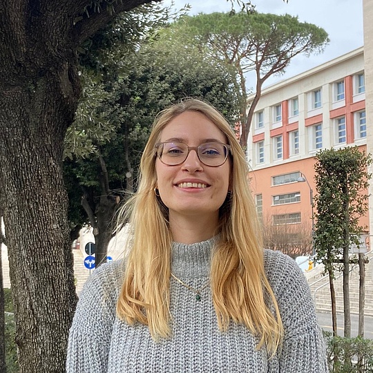 Blonde Frau in grauem Strickpullover steht zwischen schattigen Bäumen vor einem hellen Universitätsgebäude mit breiter Treppe und hohen Säulen.