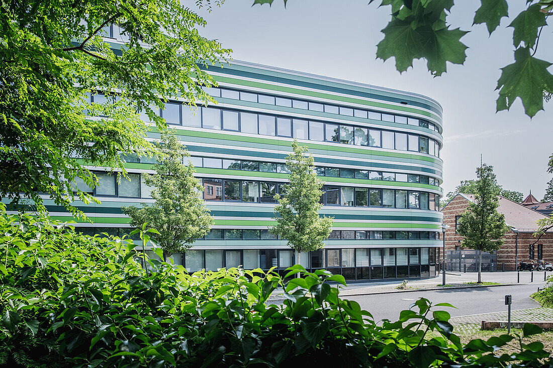 Rhoda-Erdmann-Haus mit vielen Fenstern, runder Gebäudeform und einer breiten Straße davor. Zu sehen sind auch viele Bäume und Gebüsche rund um das Gebäude.
