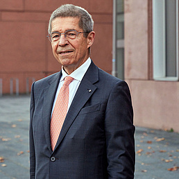 Ein Mann mit Brille, grauem Haar, in Anzug und Krawatte, steht vor einem Gebäude.