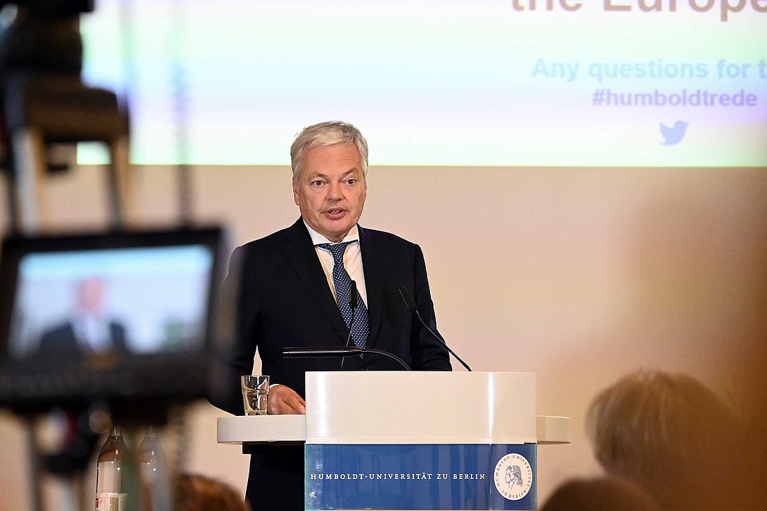 Didier Reynders am Pult. Eine Kamera ist auf ihr gerichtet, im Hintergrund ist eine Präsentation zu sehen.