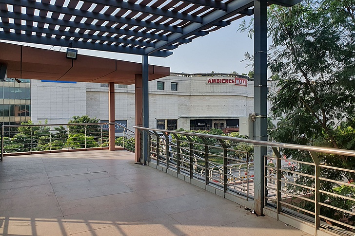 Blick von einer erhöhten Terrasse mit Holzboden und Metallgeländern auf das weiße Mehrgeschossgebäude des Ambiance Mall, umgeben von Bäumen und Pflanzen unter blauem Himmel.