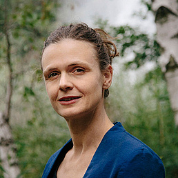 Aileen Edele trägt eine royalblaue Jacke und ihre Haare sind in einem Dutt hochgesteckt. Im Hintergrund sind Birkenbäume und Gräser zu sehen. Sie blickt freundlich die Kamera.