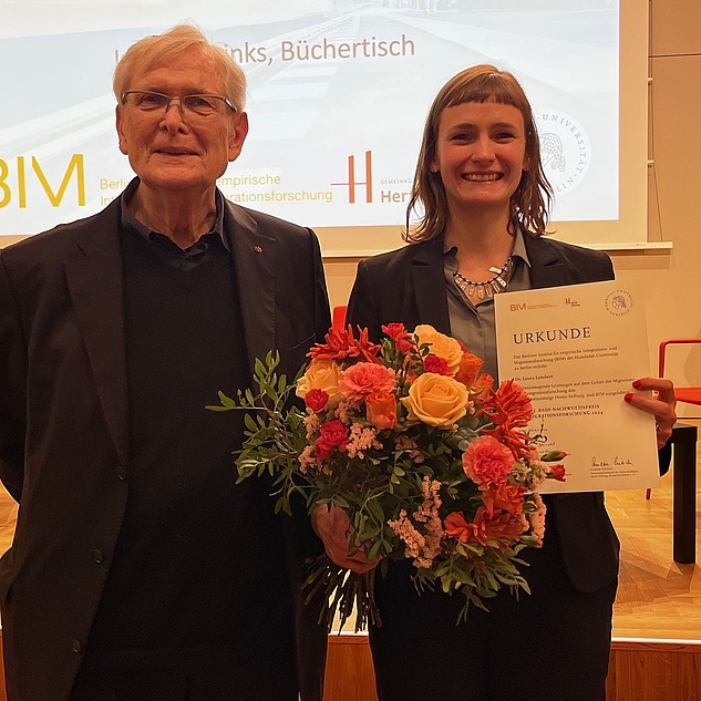 Dr. Laura Lambert mit Klaus J. Bade im Senatssaal der HU. Beide stehen nebeneinander, sie hat einen Blumenstrauß und eine Urkunde in der Hand. Im Hintergrund ist eine Präsentation zu sehen.