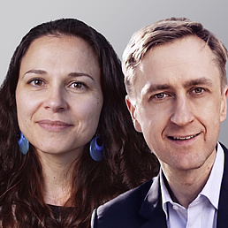 Eine Frau mit dunklen Locken und blauen Ohrringen sowie ein Mann im Anzug lächeln vor grauem Hintergrund.