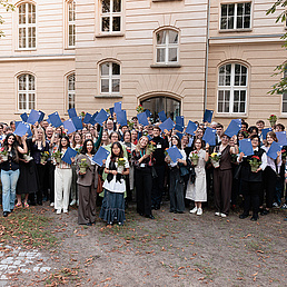 Stipendiat*innen des Deutschlandstipendiums stehen als Gruppe vor einem Gebäude und tragen sommerliche Kleidung. In den Händen halten sie ihre Urkunden hoch.